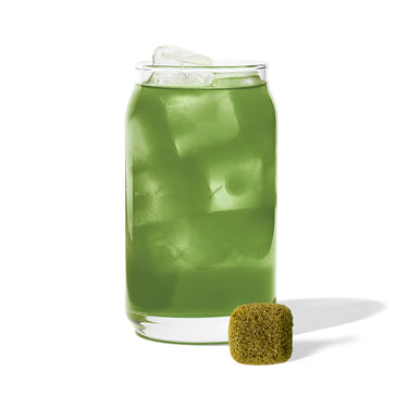 A clear glass filled with ice and a green Lavender Matcha Tea Drop – Calm Energy in Every Sip 💜🍵 sits on a white surface. In front of the glass is a small, square greenish-brown cube.